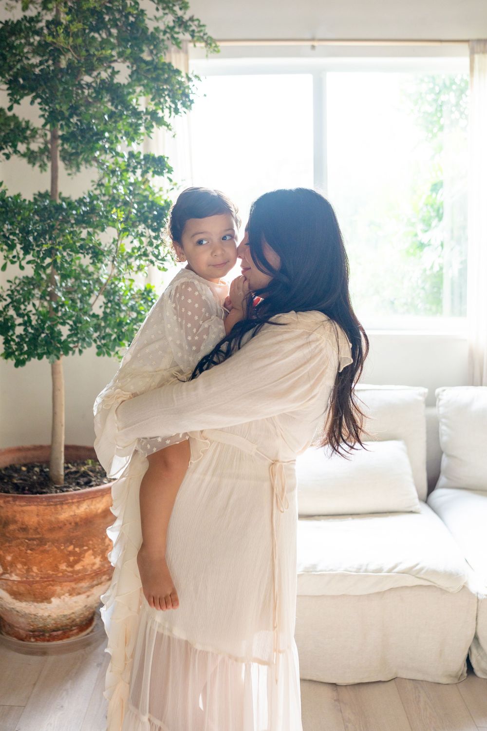 Two figures embrace in a bright white room with a potted plant beside them.