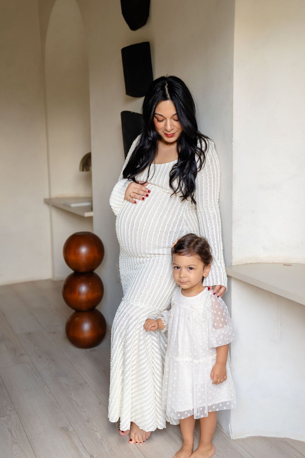 A mother and daughter wearing matching white dresses pose together in a minimalist interior setting.