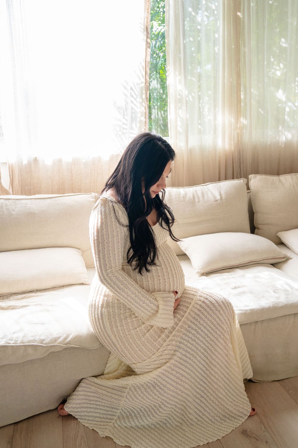 Someone in a cream knit dress sits gracefully on a white couch near sheer curtains.