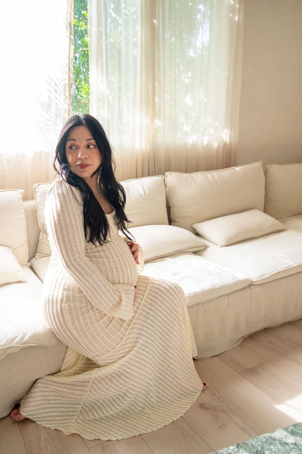 A person in a cozy cream knit sweater dress poses on a white couch in a sunlit room.