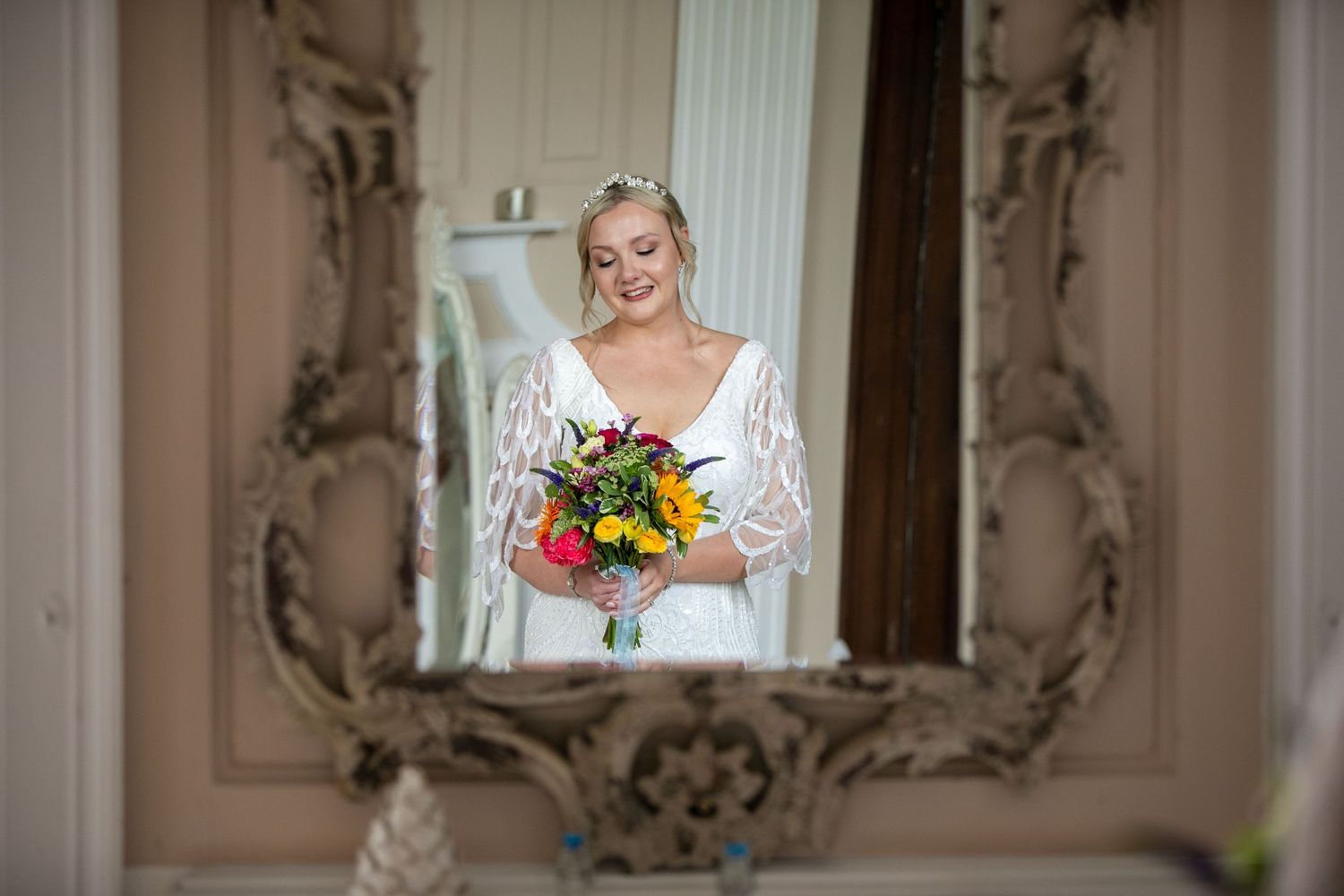 Bride in a mirror reflection, wearing a white gown with lace sleeves, holds a vibrant bouquet.