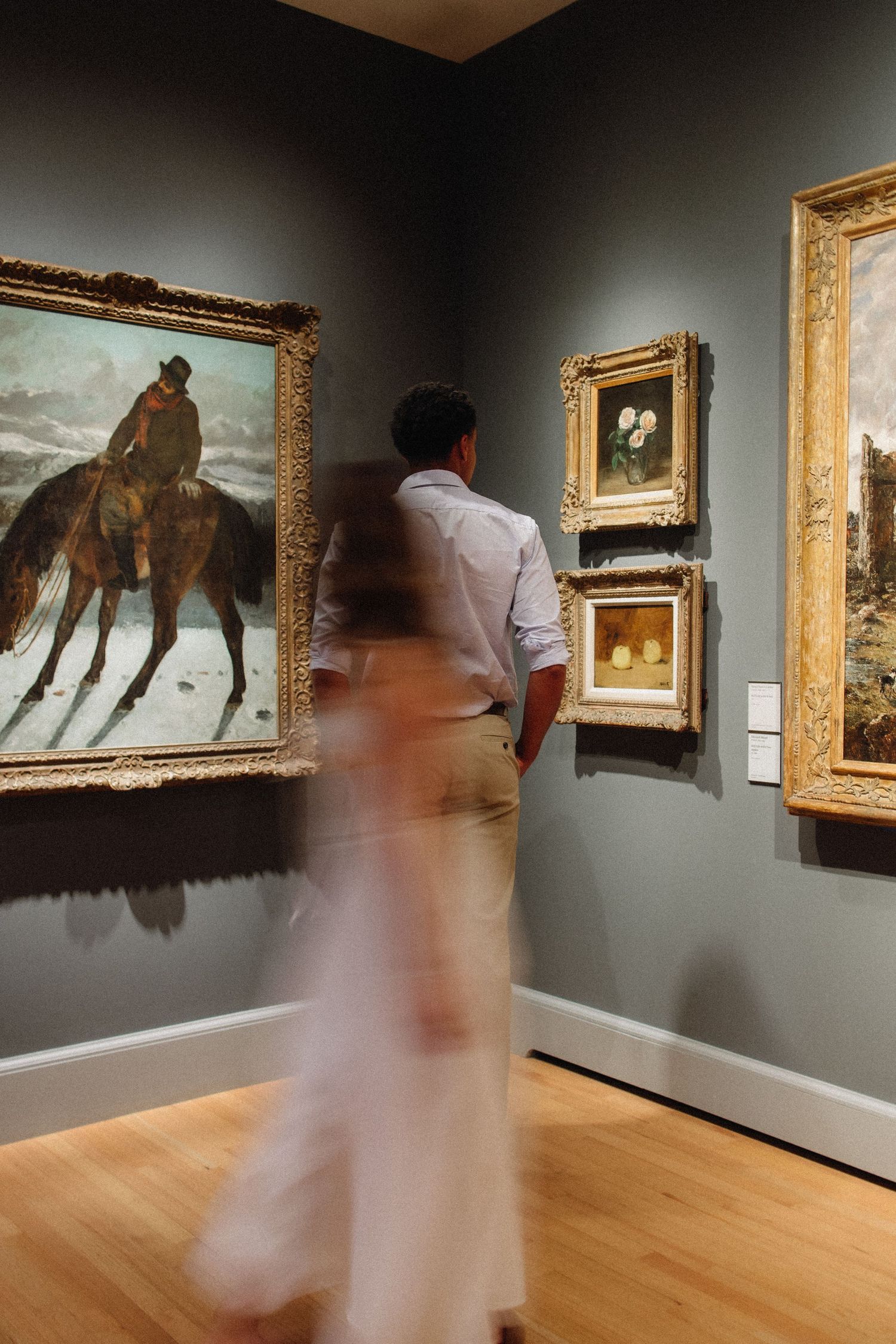 Motion-blurred visitors observe artwork displayed on gray walls in a museum gallery with wooden floors.