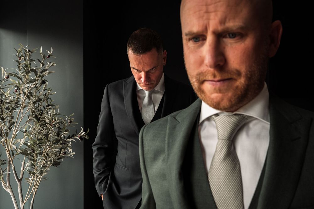 Two men in formal suits standing in dramatic lighting against a wall with decorative plants.