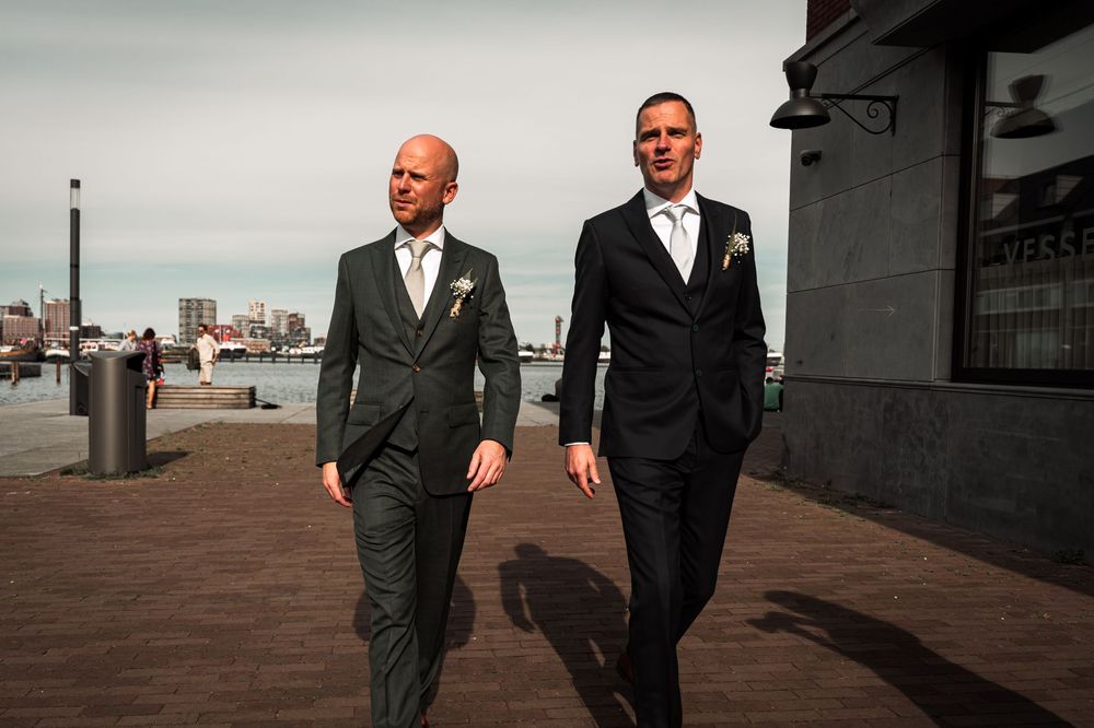 Two men in dark suits walk along an outdoor deck with city views in the background.