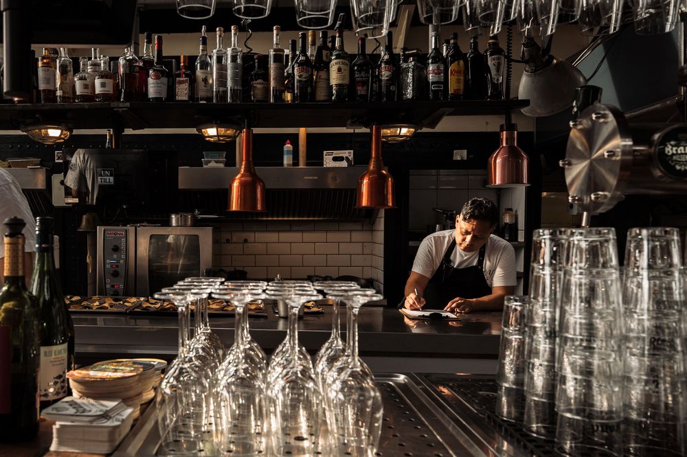 Bartender working behind counter with rows of glasses and bottles in moody lighting.
