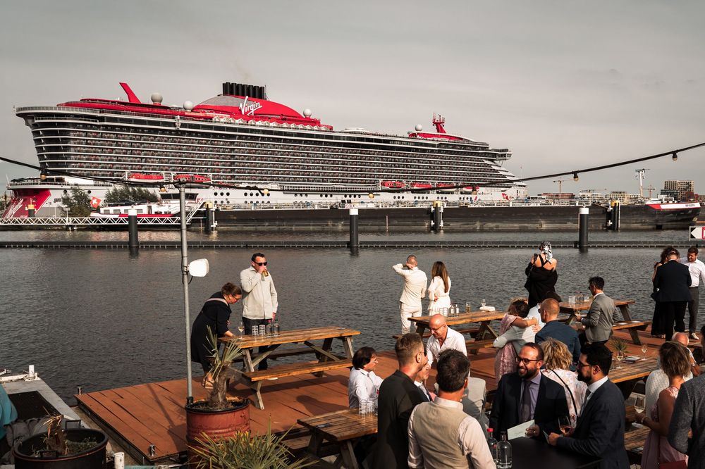 People gather at a waterfront deck overlooking a massive red and white cruise ship docked at port.