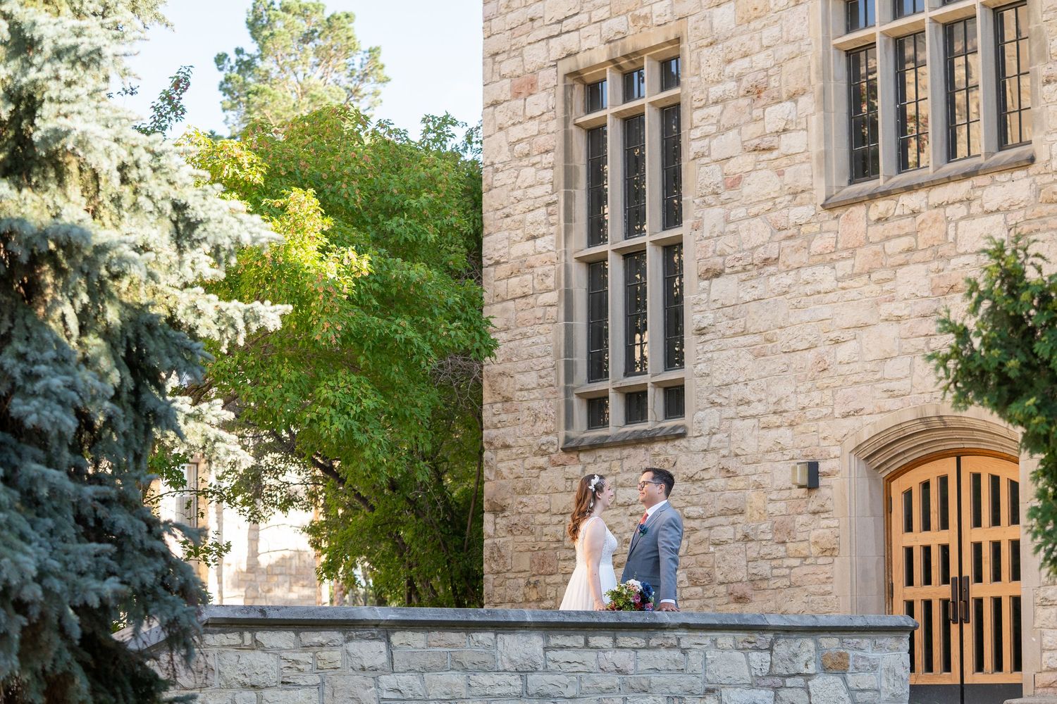 A couple stands on a stone wall outside a Gothic-style building with tall windows and stone facade.