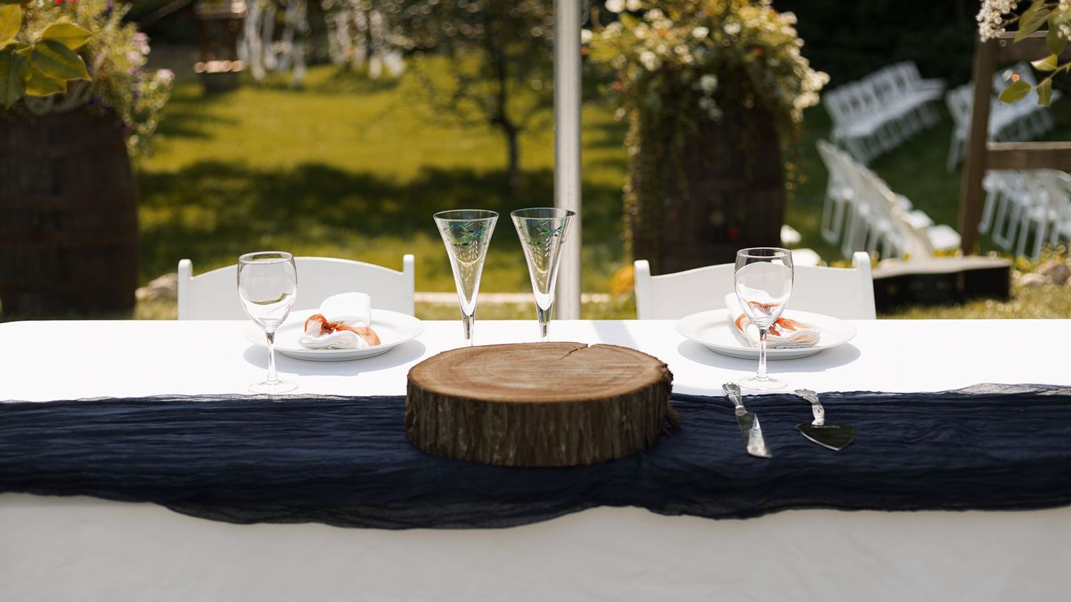 Elegant outdoor wedding reception table set with white chairs, glassware, and a rustic wood centerpiece.