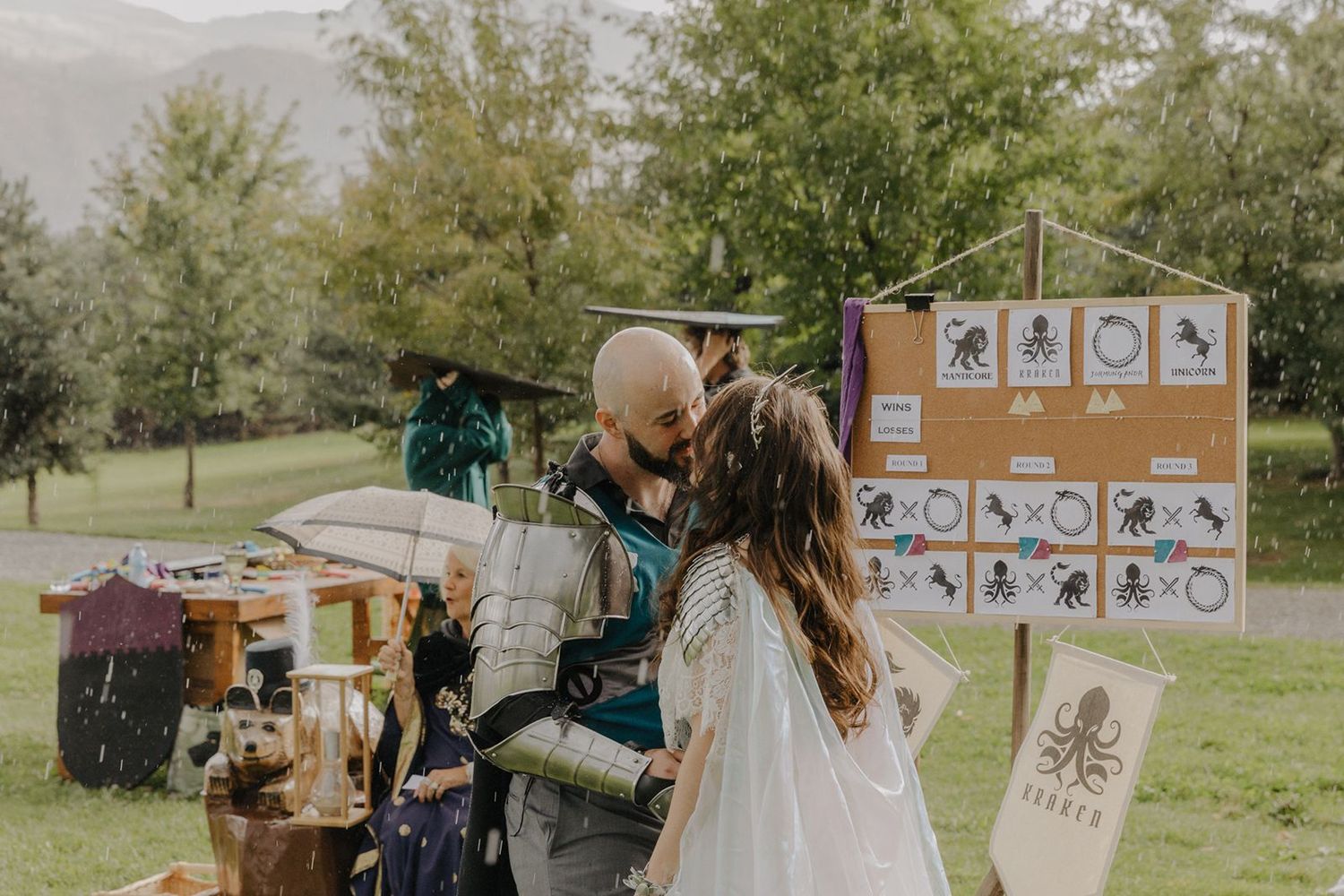 Couple sharing a kiss in the rain at a fantasy-themed outdoor wedding with decorative banners.