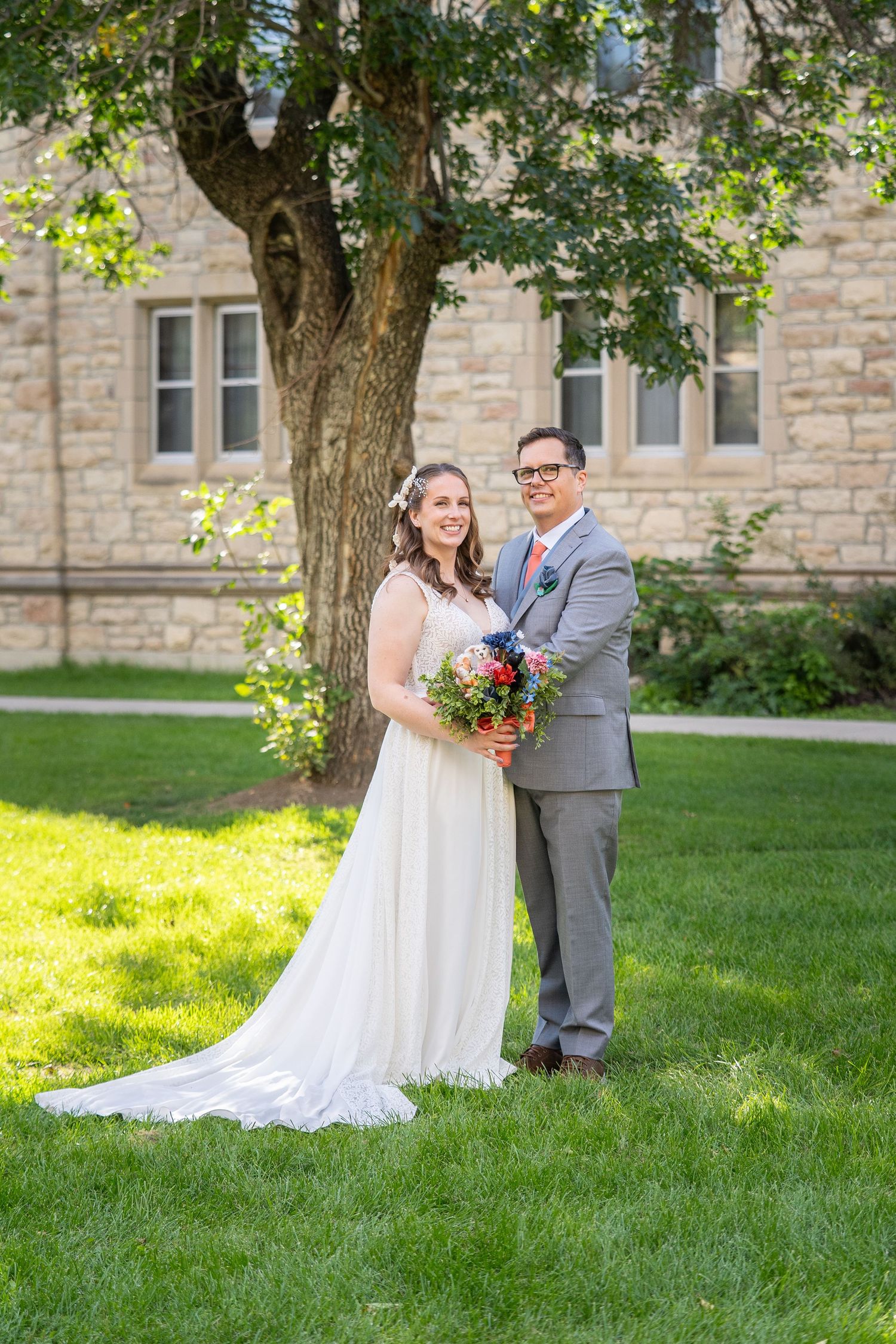 A couple in wedding attire poses together on a sunny lawn in front of a stone building with a large tree.
