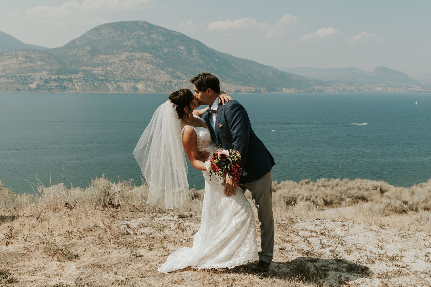 Bride and groom sharing a kiss on hilltop with stunning blue lake and mountain scenery behind them.