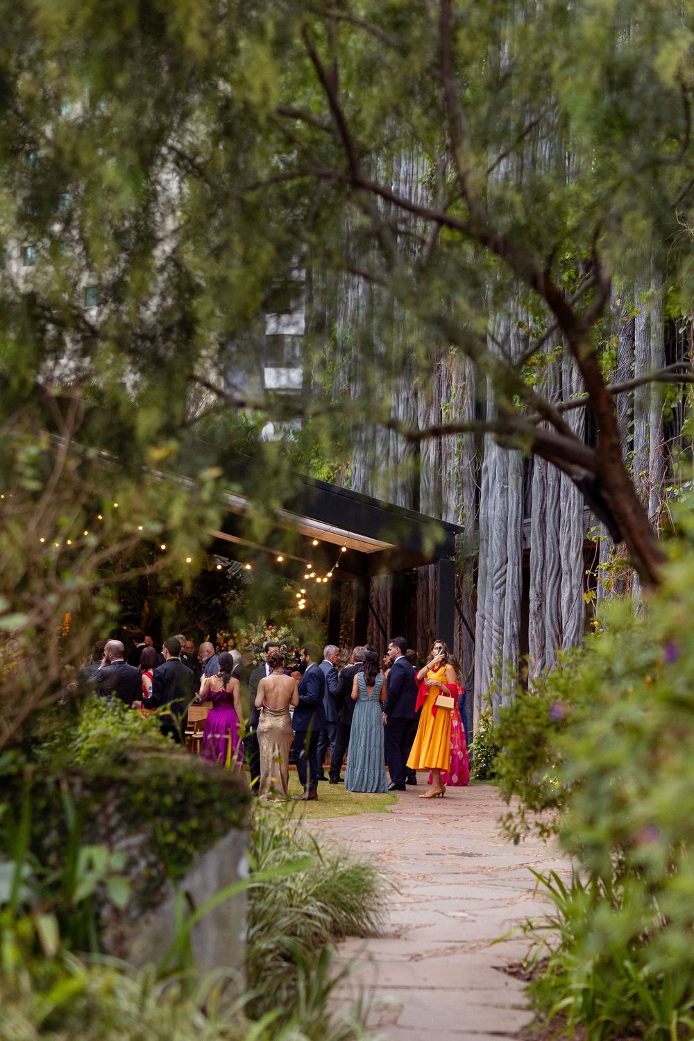 Wedding guests gather on a tree-lined outdoor path decorated with string lights for an evening celebration.