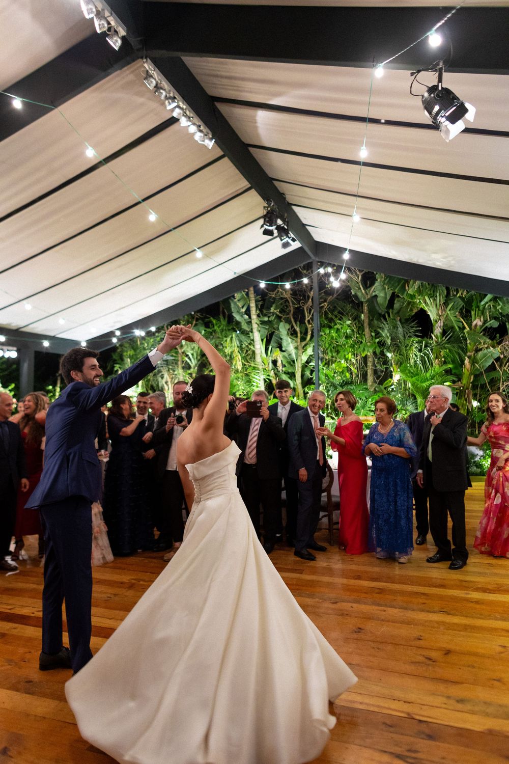 A bride and groom share their first dance surrounded by guests in an elegant reception venue with wood floors.