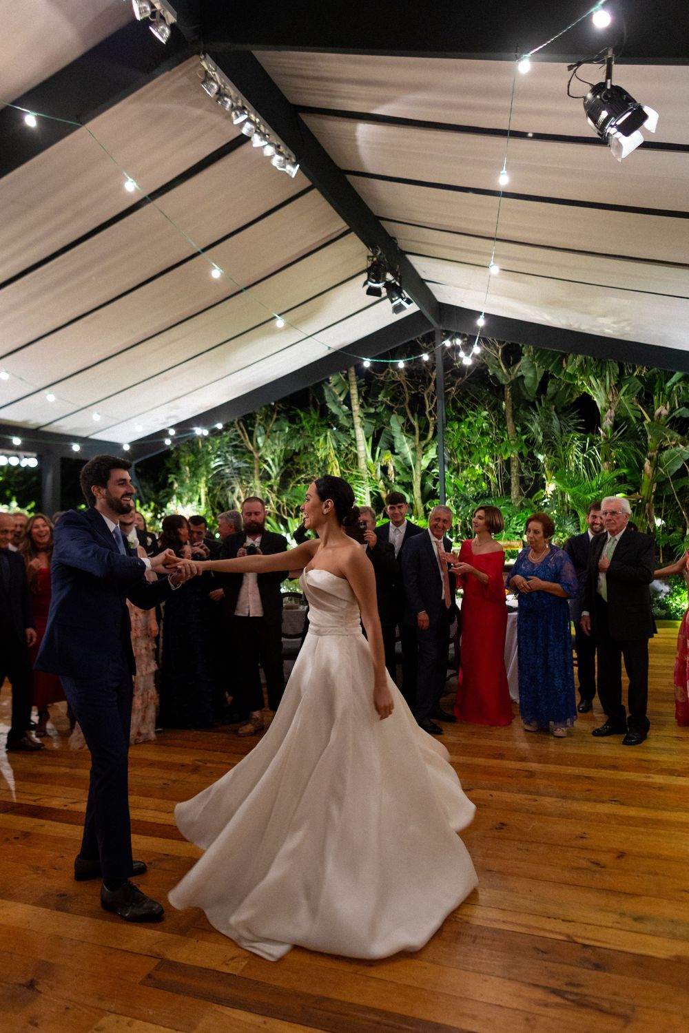A couple performs their first dance at a wedding reception on a wooden dance floor under a tented venue with greenery.