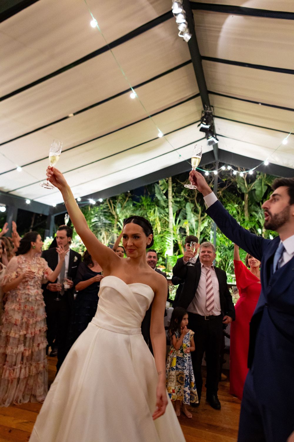 Wedding reception guests dancing and celebrating on a wooden dance floor under track lighting with greenery in background.