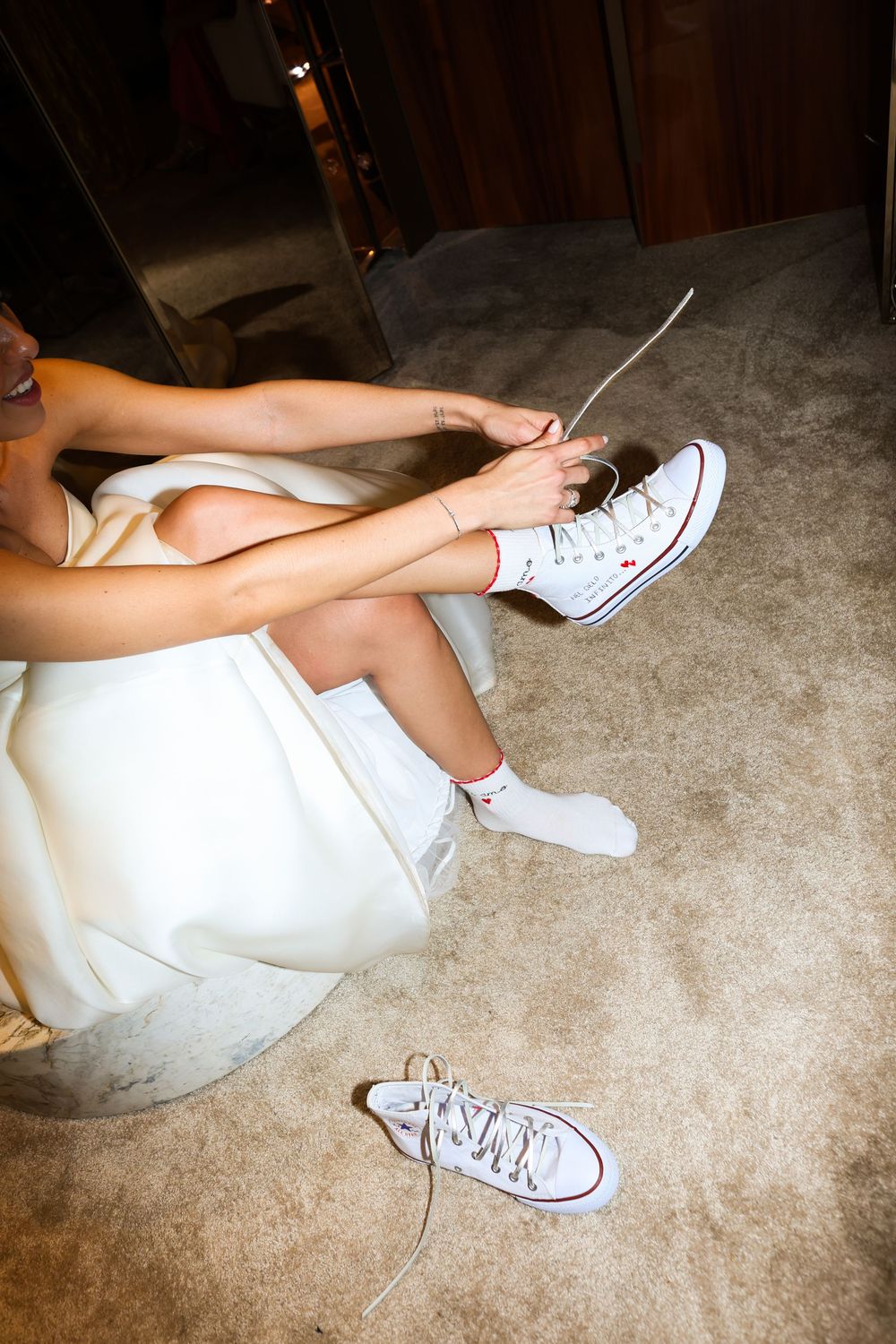 Close-up of white Converse sneakers worn with a wedding dress on a beige carpet.