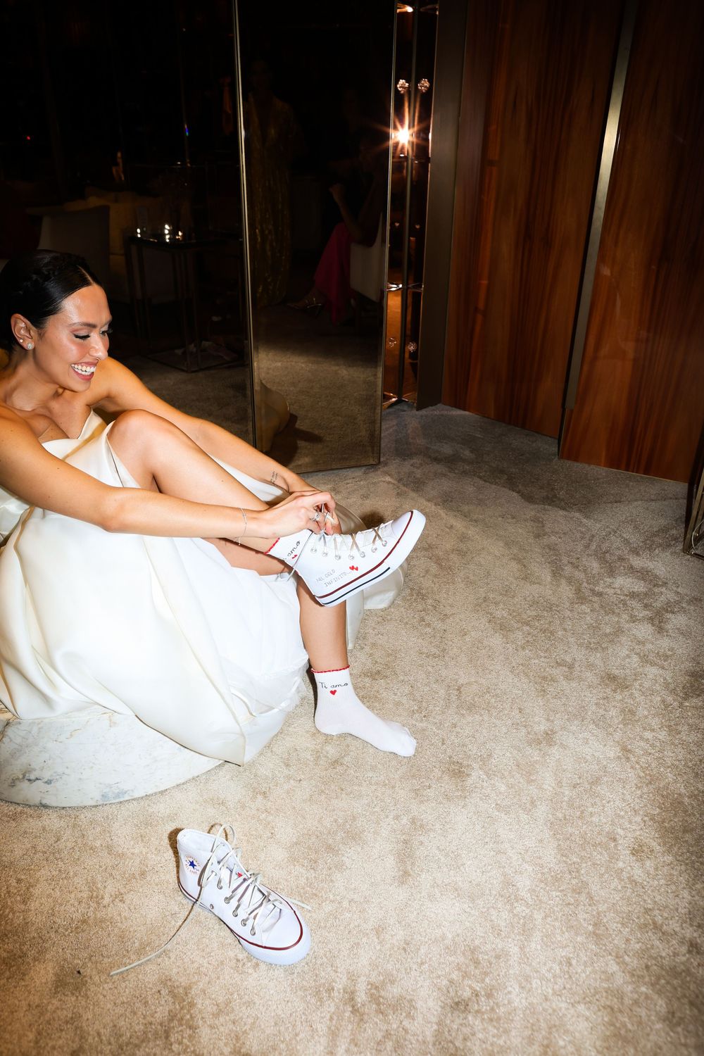 Person in white dress sitting on carpet putting on white Converse sneakers.