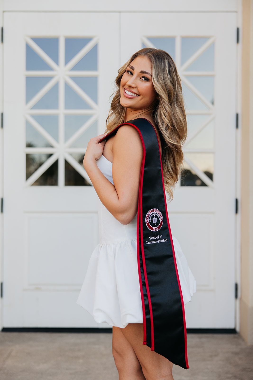 Graduate in white dress and graduation stole poses for photos in front of white garage doors.