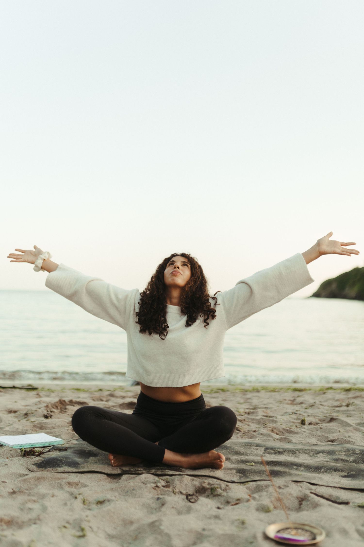 A person sits in lotus position with arms outstretched on a sandy beach at sunset.