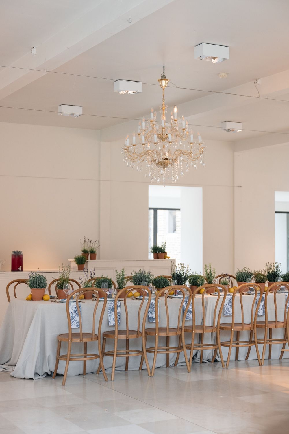 Elegant banquet hall setup with long tables, wooden chairs, and crystal chandeliers in a bright white space.