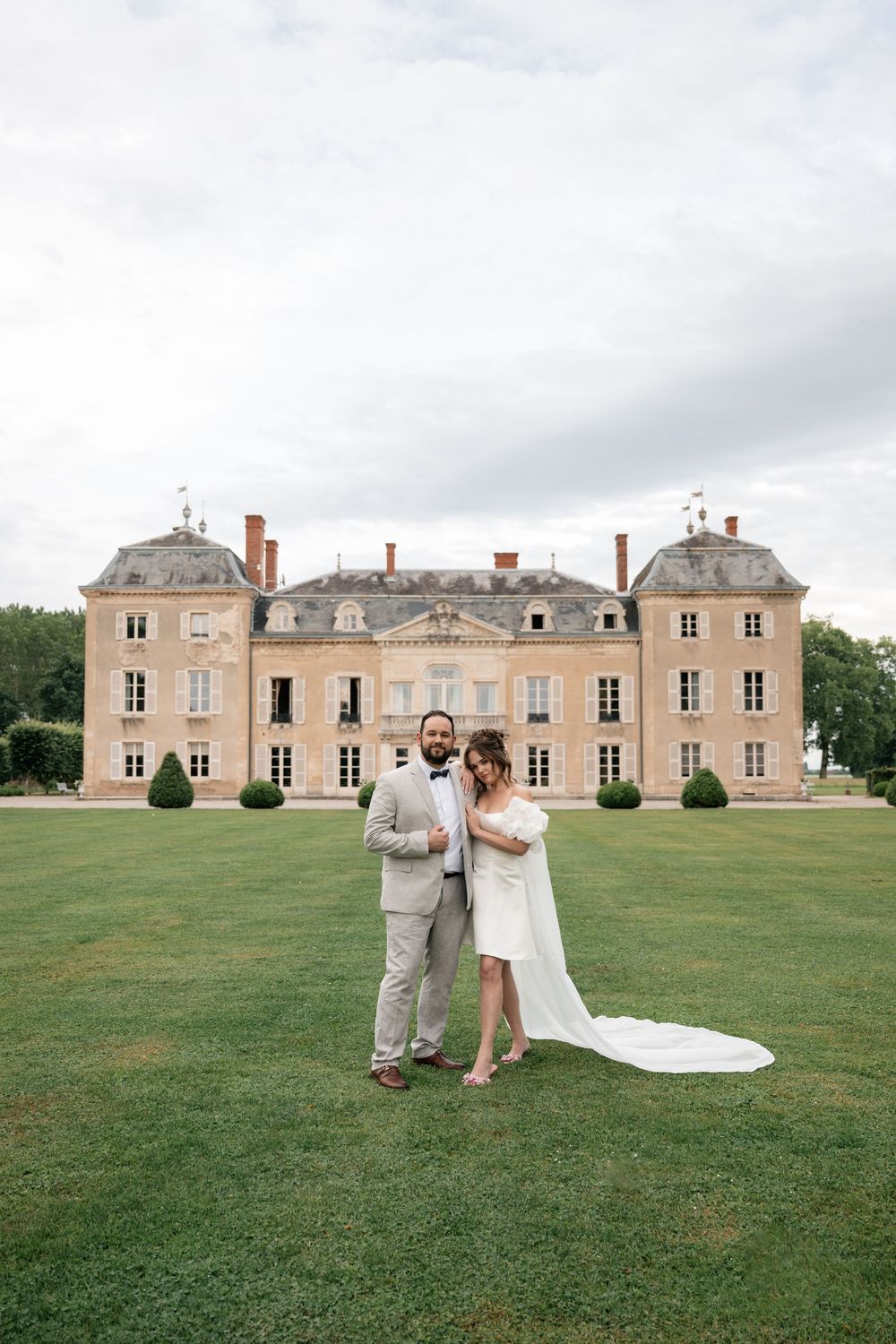 A wedding couple poses on the lawn in front of an elegant French chateau with grey stone facade and dormer windows.