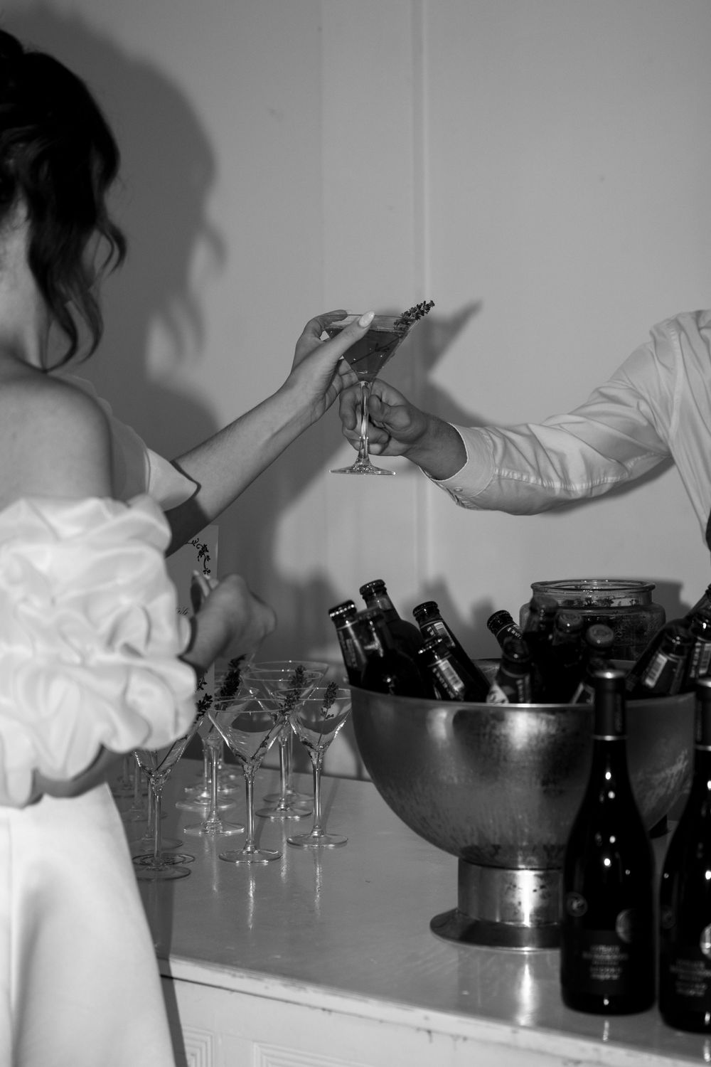 Black and white image of wine glasses and bottles being served at an indoor gathering.