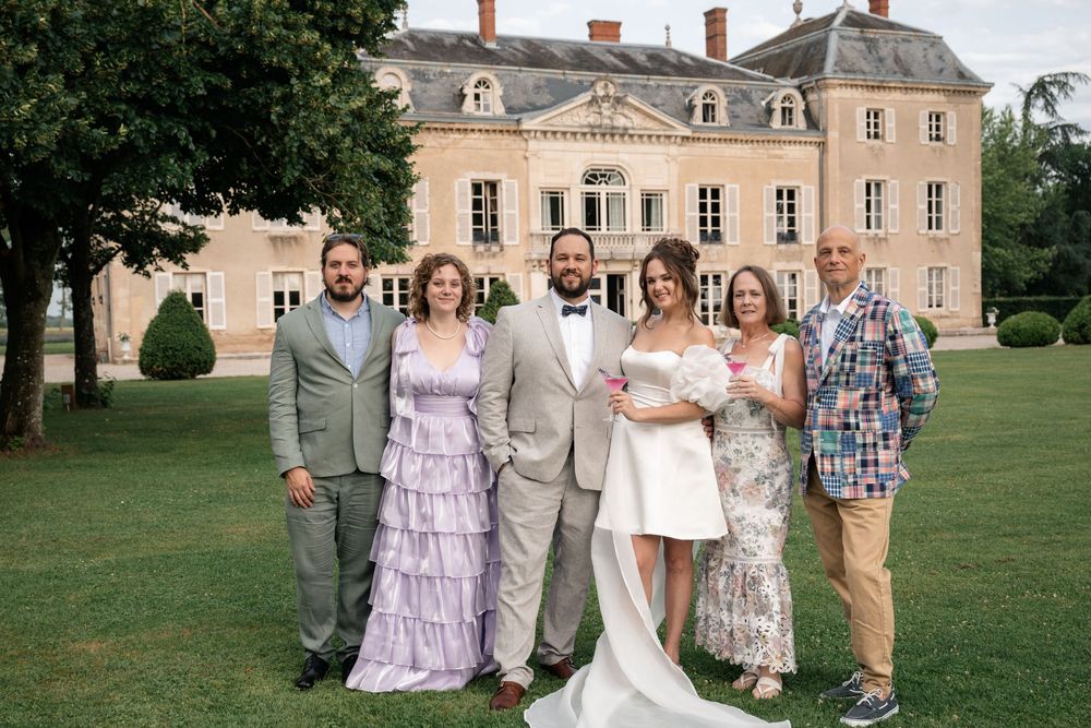 Wedding party poses for formal portraits on the lawn of an elegant chateau with pink stone exterior and manicured grounds.