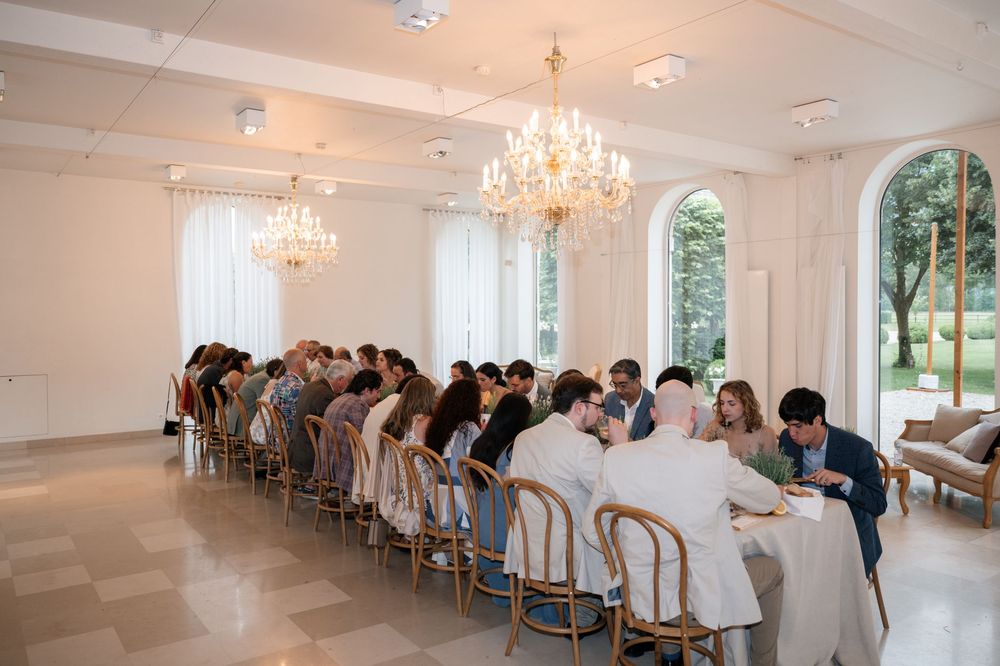 Wedding guests seated in elegant white chairs face towards the front of a bright room with crystal chandeliers.
