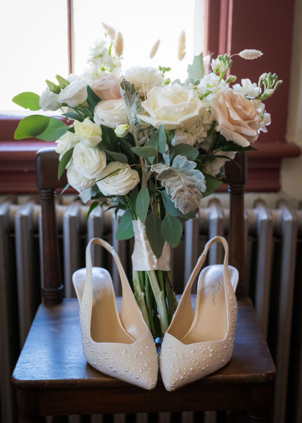 Bridal bouquet of white and blush roses with eucalyptus beside pearl-embellished ivory slingback wedding heels on wooden chair.