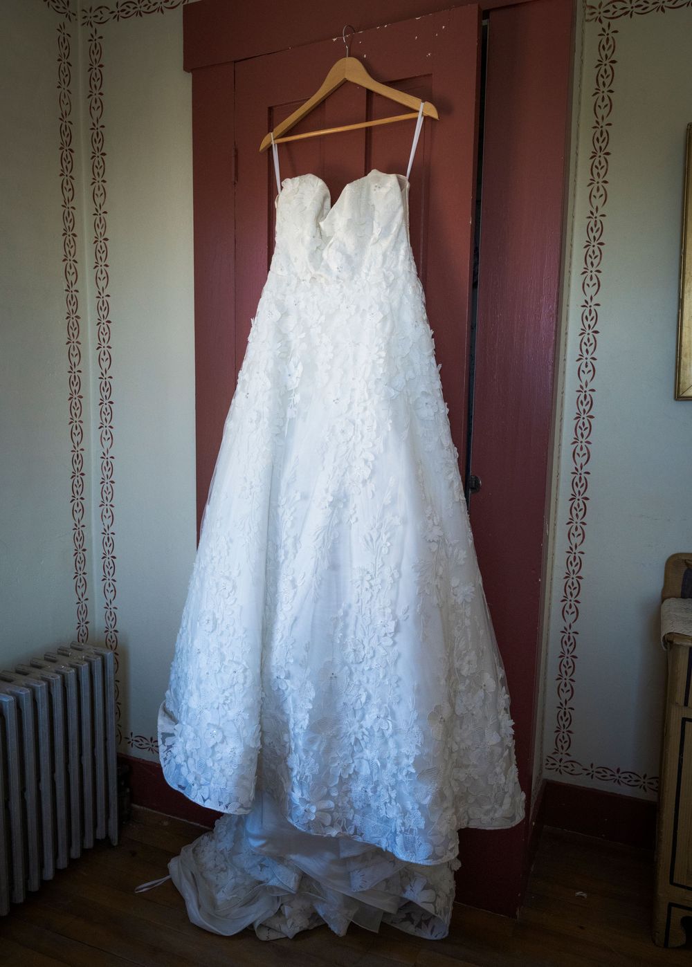 Elegant strapless white lace ball gown wedding dress hanging on a wooden hanger against a rustic door.