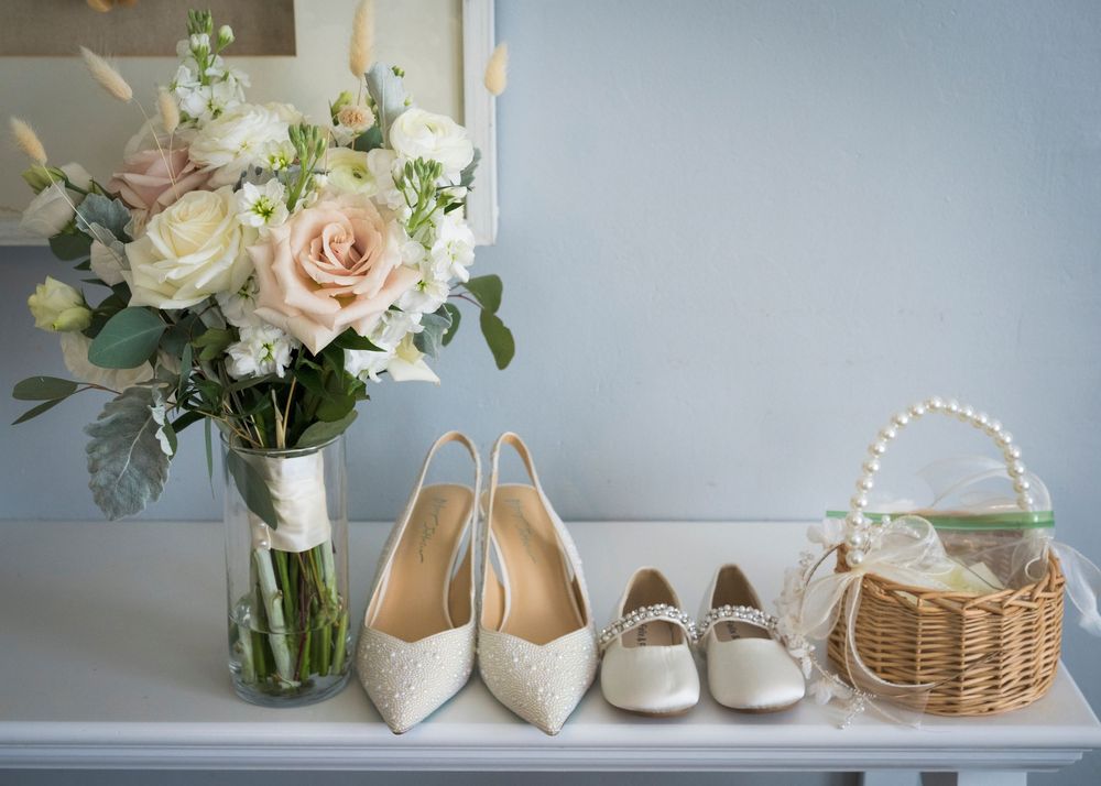 Wedding flat lay with blush rose bouquet, ivory heels, flower girl shoes, and pearl basket on white shelf.