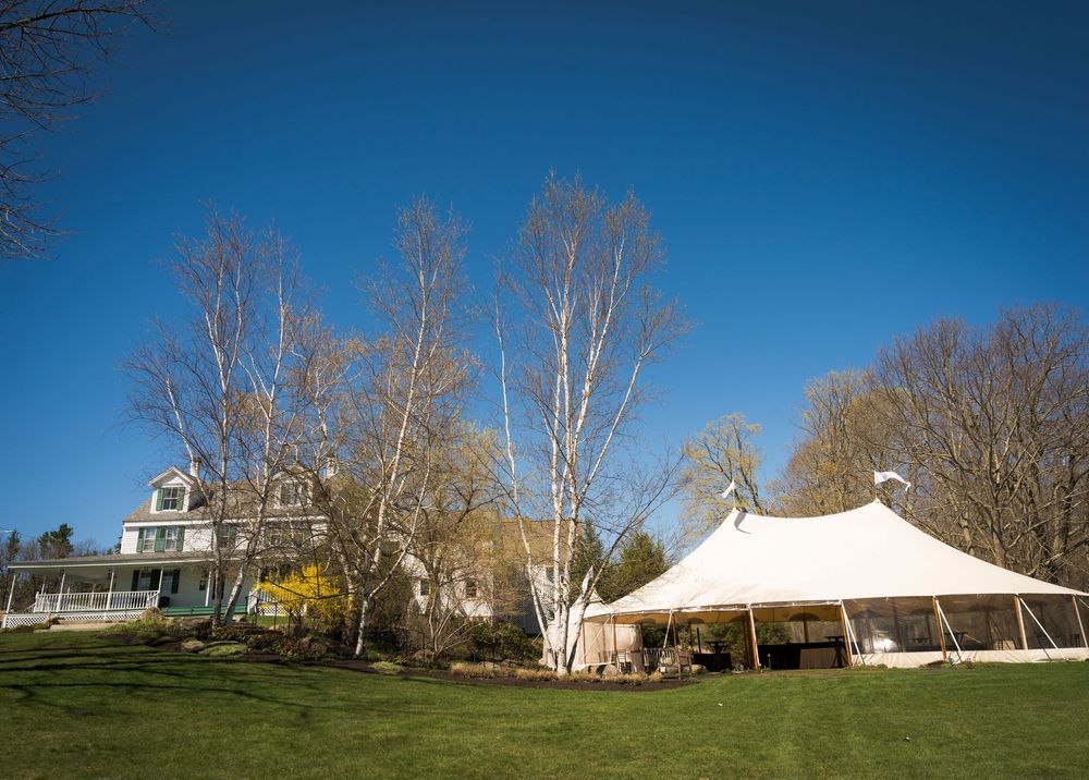 Elegant wedding venue with white tent and historic farmhouse on lush green lawn under bright blue sky.