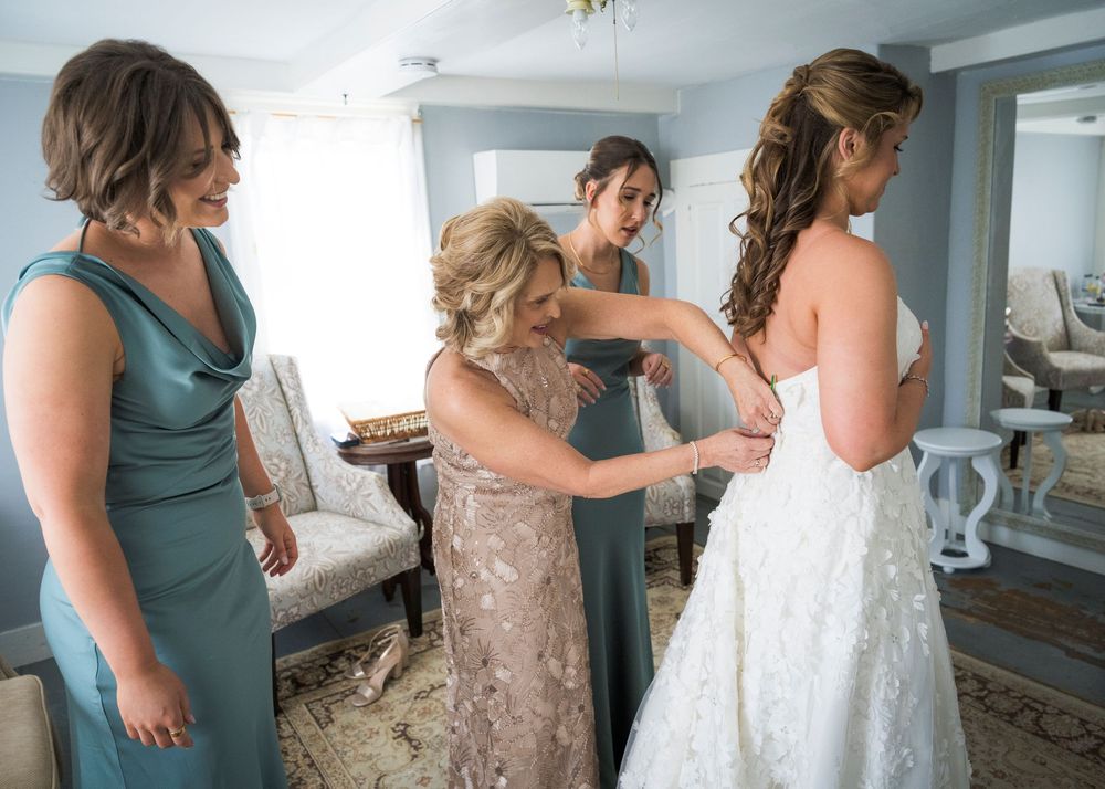 Mother and bridesmaids in teal gowns help fasten the bride's lace wedding dress before the ceremony.