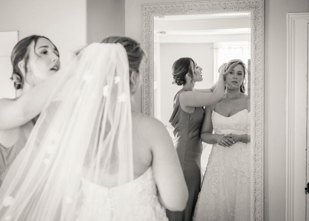 Bridesmaids help bride with her veil while she admires her wedding gown reflection in a large mirror.