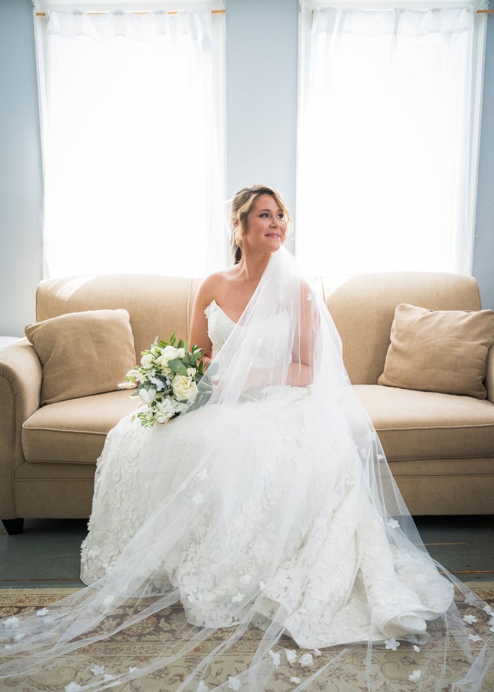 Bride in lace wedding gown and veil sits on beige sofa holding white floral bouquet by bright windows.