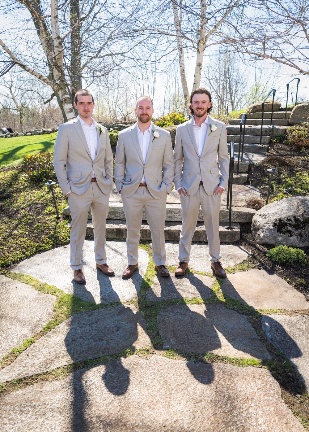 Groom and two groomsmen in matching tan suits posing outdoors on a stone patio.