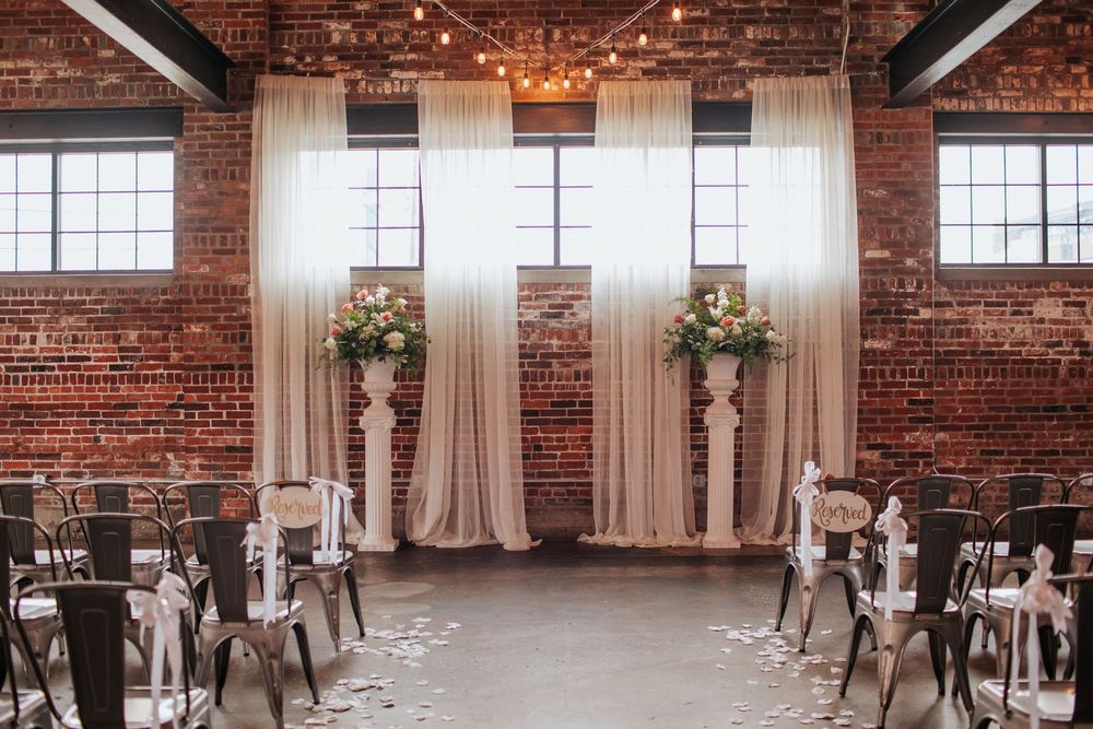 Industrial chic wedding venue with exposed brick walls, tall windows, and elegant white drapes creating a romantic atmosphere.