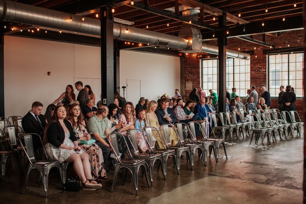Wedding guests seated in rows of gray chairs in an industrial-style venue with exposed brick walls and string lights.