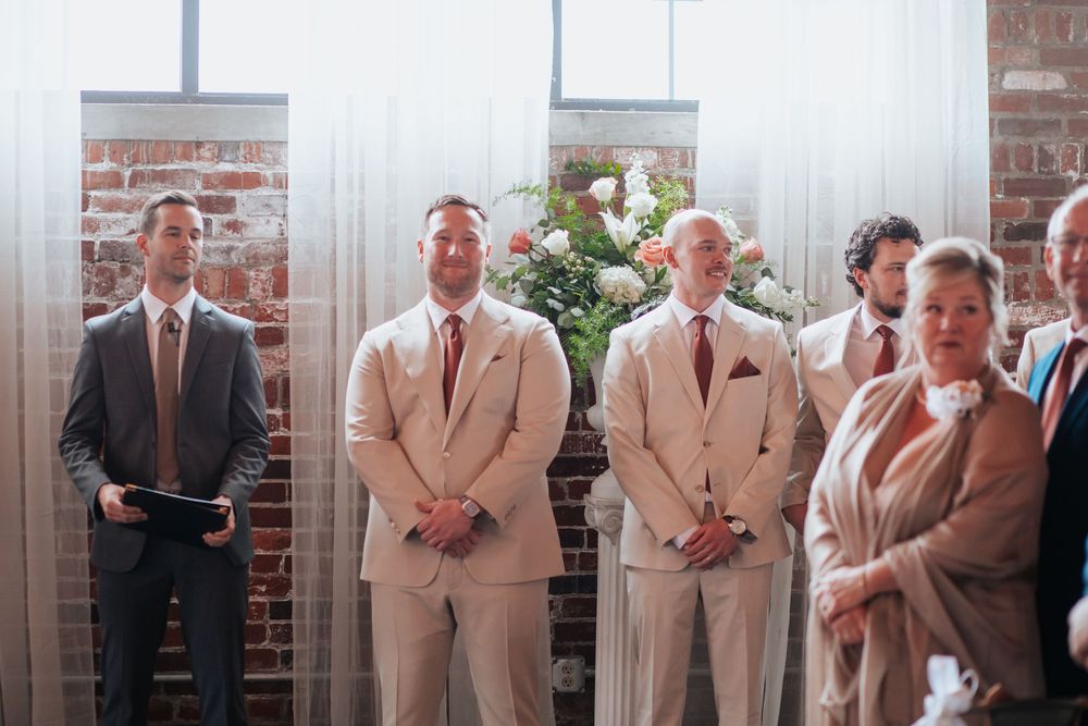Wedding party members standing in a line against a brick wall wearing light beige and neutral colored attire.
