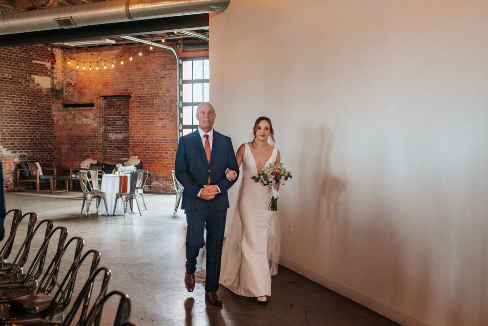 A couple walks arm in arm down an industrial-style hallway with exposed brick walls and string lights at a wedding venue.