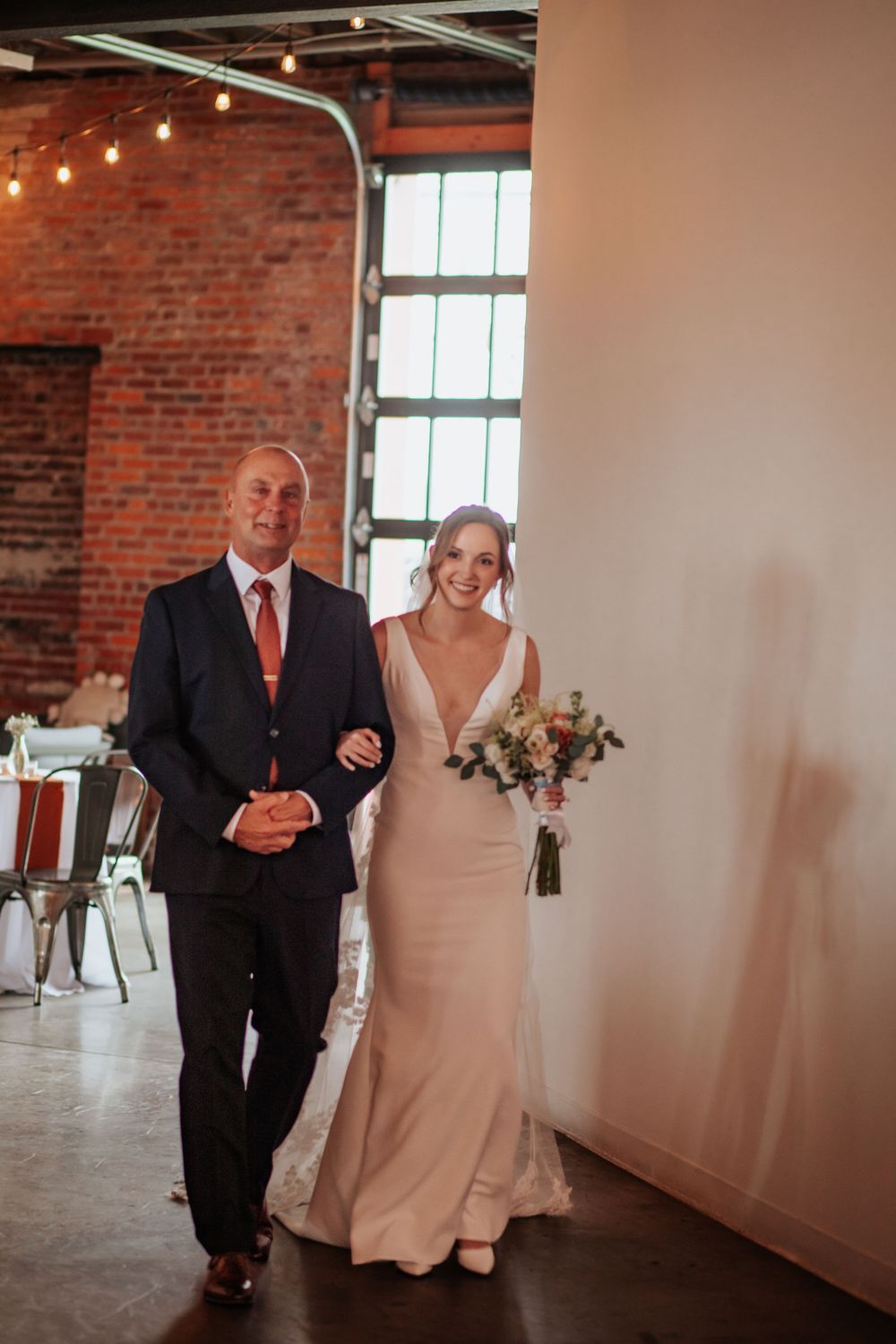 Wedding couple walks down an aisle in an industrial brick venue with string lights overhead.