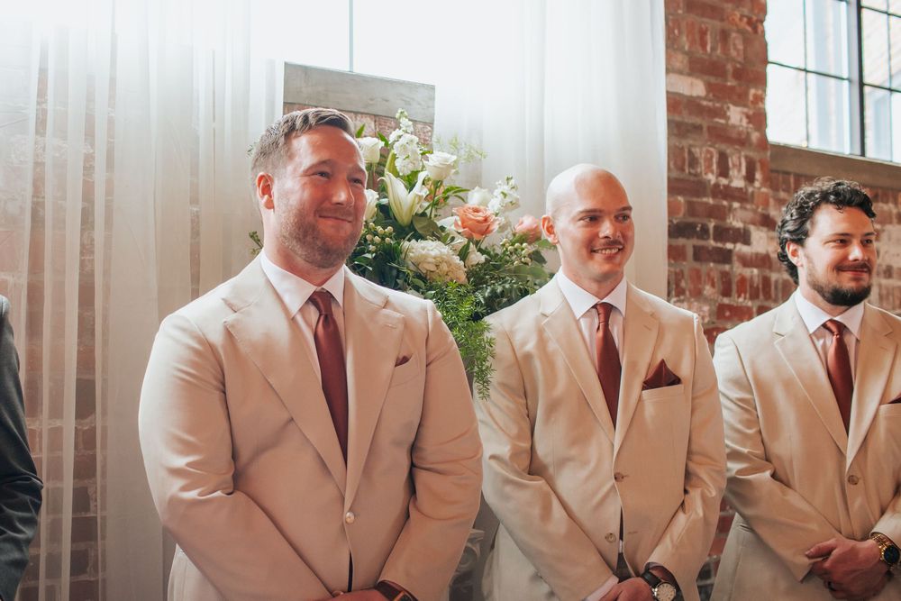 Groomsmen in beige suits and brown ties stand in line at an indoor wedding venue with exposed brick walls.
