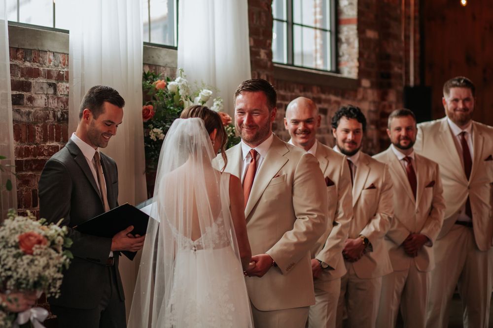 A wedding ceremony in progress with groomsmen in beige suits and a bride in a white veil at an indoor venue.