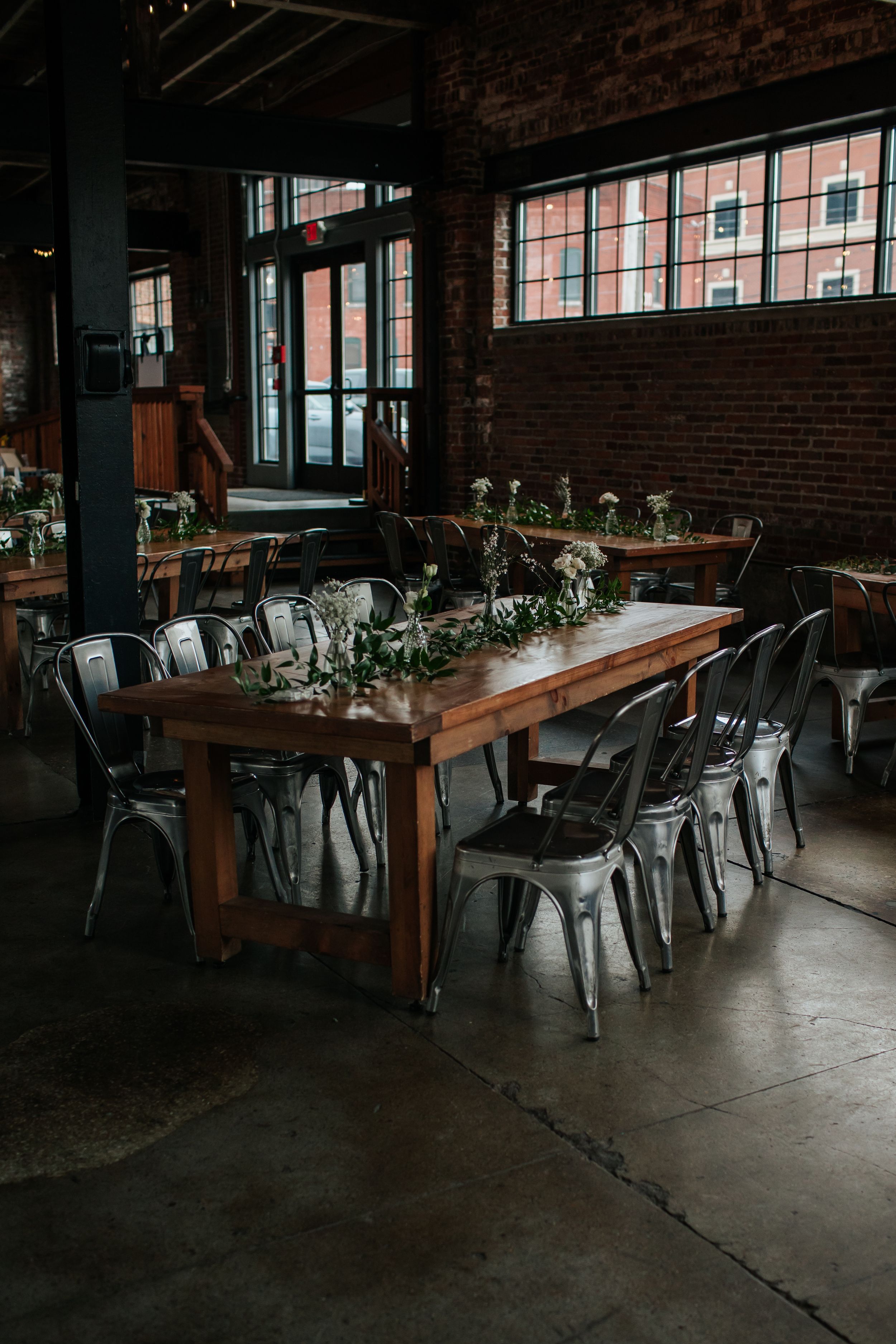 Long wooden tables with metal chairs are arranged in an industrial-style event space with exposed brick walls and greenery.