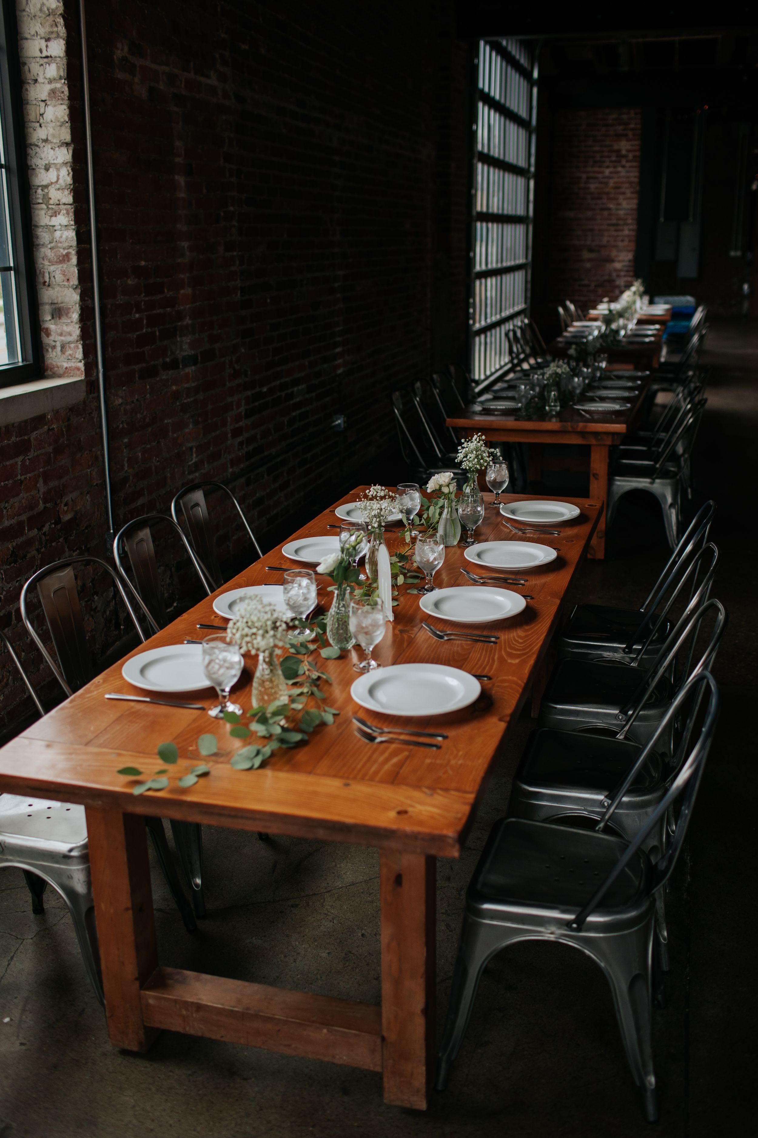 Long wooden dining table with black chairs and white place settings in a rustic industrial space.