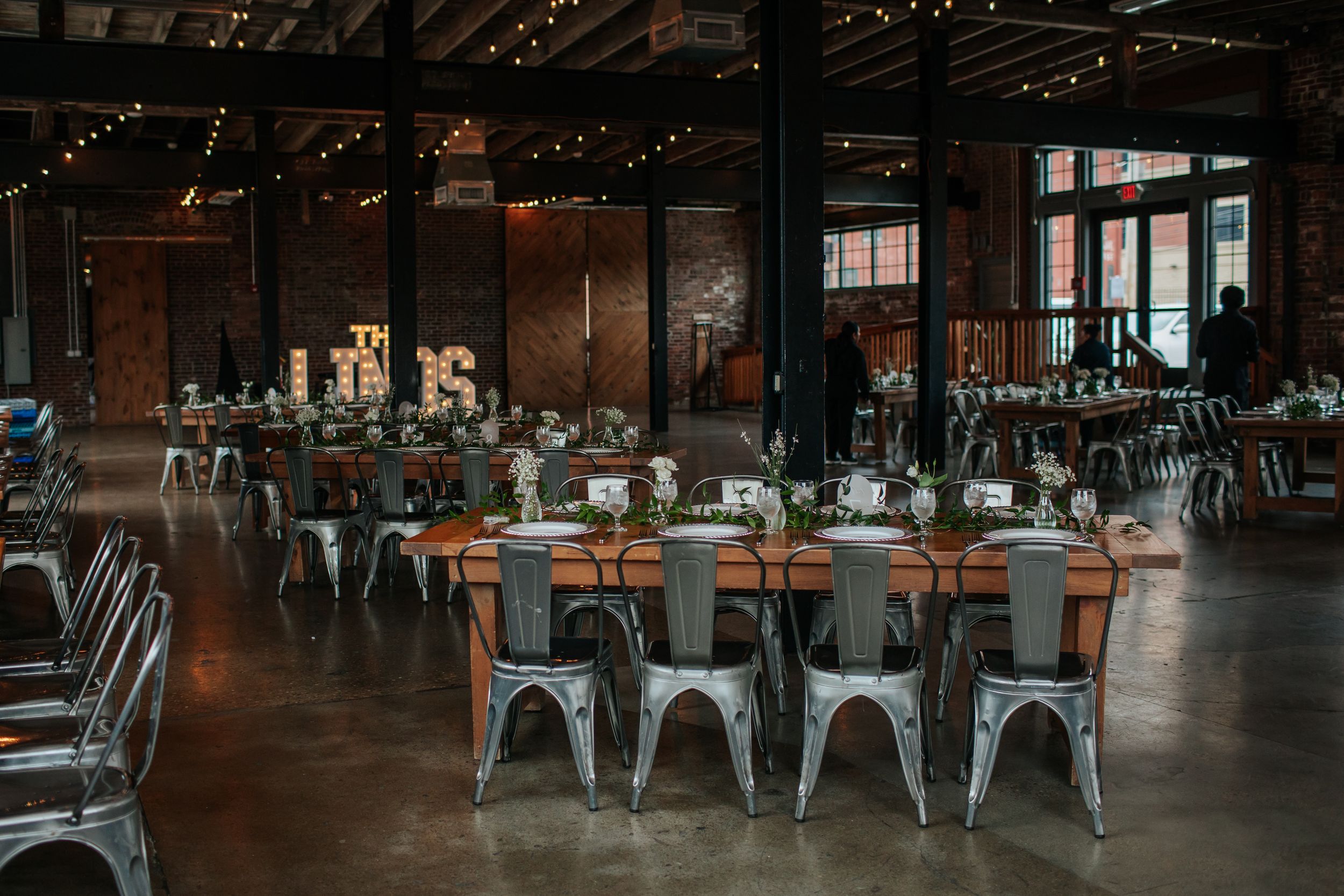 A dark industrial-style restaurant interior with metal chairs and exposed ceiling beams illuminated by warm lighting.