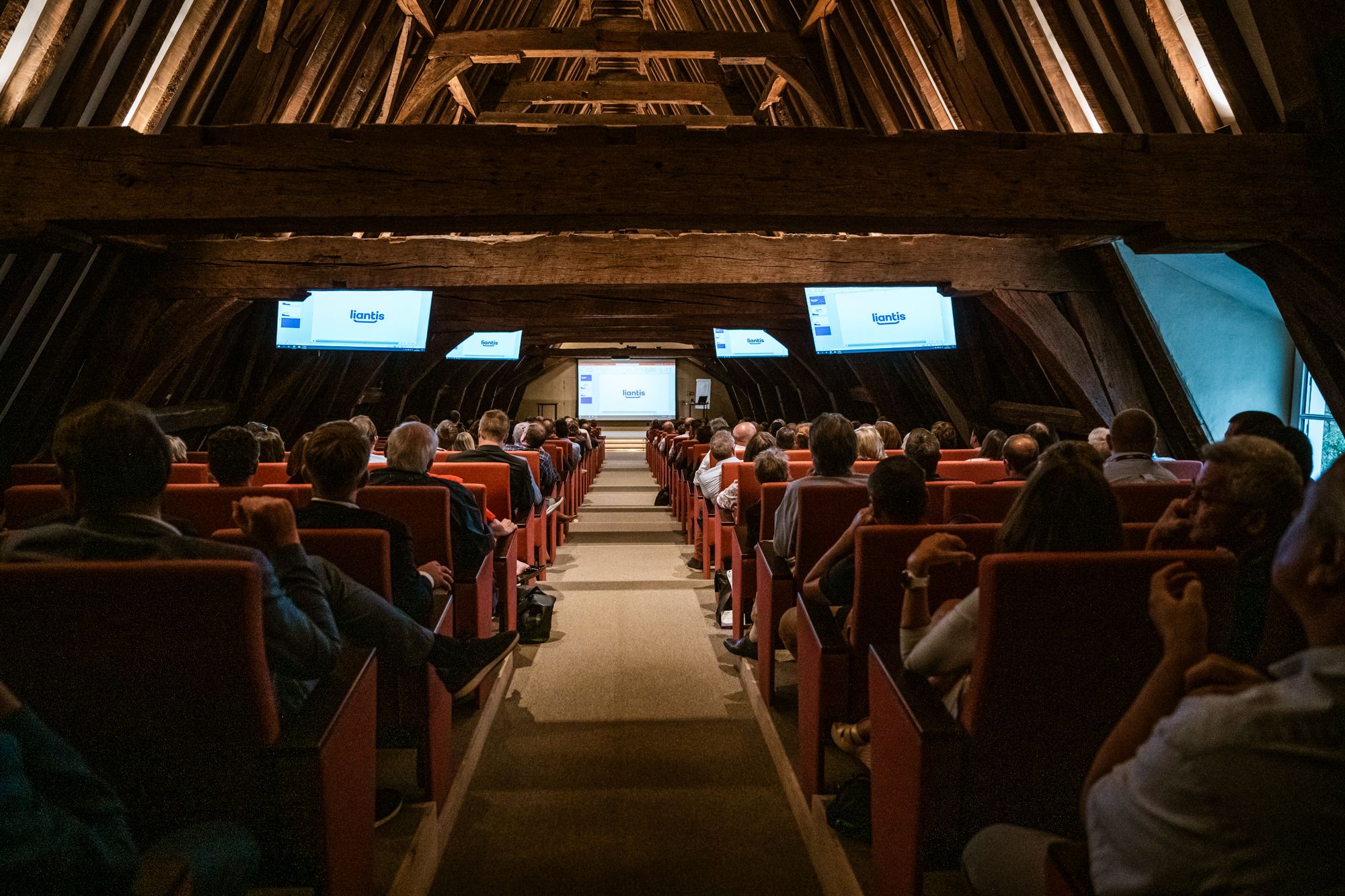Een volle conferentiezaal met houten dakconstructie, waar deelnemers aandachtig naar een Liantis-presentatie kijken