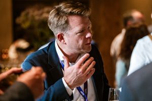 Een man in gesprek, geanimeerd pratend met een handgebaar, tijdens een netwerkevenement in een sfeervolle setting