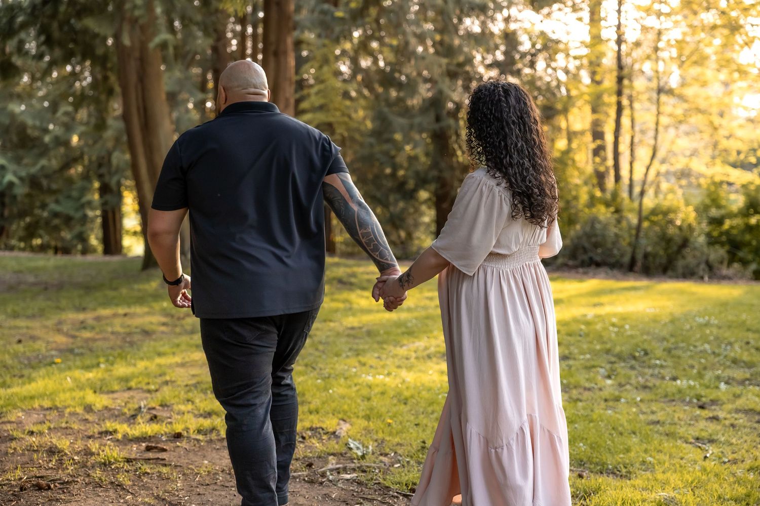 A couple holding hands while walking through a sunlit forest path in late afternoon golden light.