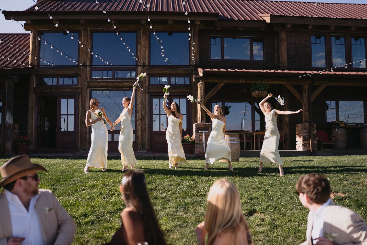 Bridesmaids in white dresses joyfully toss bouquets on green lawn outside rustic lodge wedding venue, Blame Her Ranch
